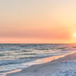 Serene Pensacola Beach sunset with gentle ocean waves rolling onto the sandy shore. The sky displays a warm gradient of orange, pink, and yellow hues as the sun nears the horizon. A distant city skyline is faintly visible in the background. The golden sunlight reflects on the water and wet sand, creating a tranquil coastal scene. A small bird is seen near the shoreline, adding a touch of life to the peaceful landscape.