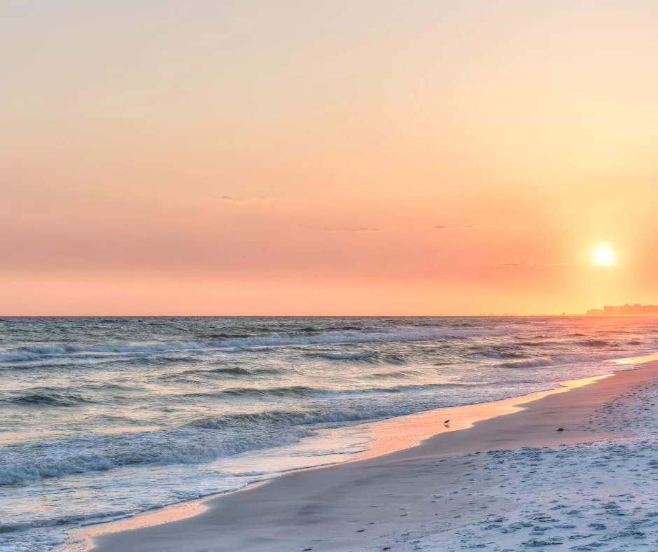 Serene Pensacola Beach sunset with gentle ocean waves rolling onto the sandy shore. The sky displays a warm gradient of orange, pink, and yellow hues as the sun nears the horizon. A distant city skyline is faintly visible in the background. The golden sunlight reflects on the water and wet sand, creating a tranquil coastal scene. A small bird is seen near the shoreline, adding a touch of life to the peaceful landscape.