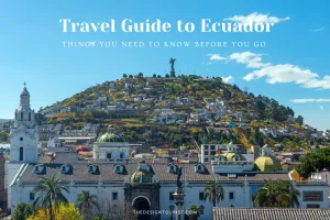 Scenic view of Quito, Ecuador with colonial architecture in the foreground and the Virgin of El Panecillo statue atop a hill surrounded by colorful hillside homes under a bright blue sky. Text overlay reads "Travel Guide to Ecuador – Things You Need to Know Before You Go."