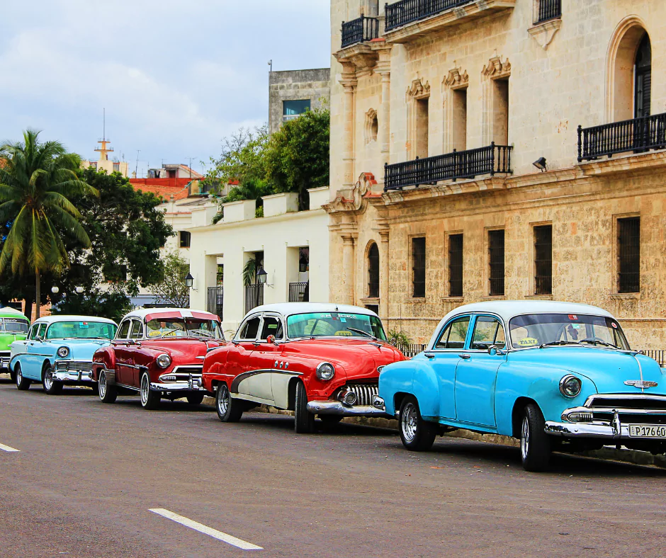 Vintage classic Cuban cars in vibrant colors parked along a street in Havana, Cuba, with colonial architecture in the background.