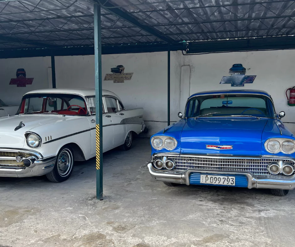 Classic Chevrolet cars, a white 1957 model and a blue 1958 model, parked in a covered garage in Cuba.