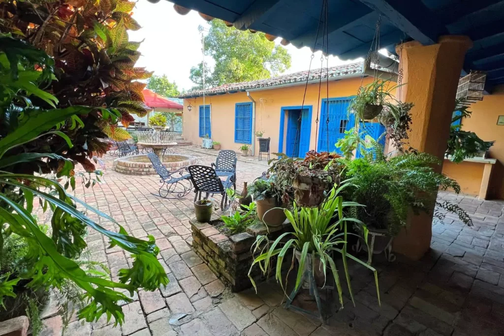 Charming courtyard of a colonial casa particular in Trinidad, Cuba, featuring colorful walls, blue window shutters, wrought iron chairs, and lush tropical plants.