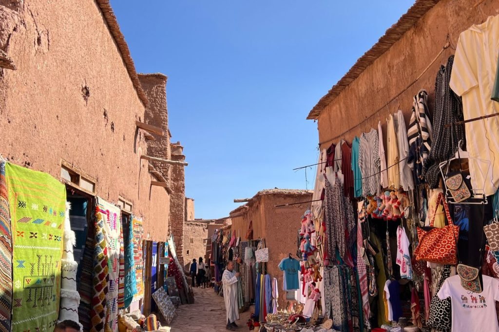 Traditional Moroccan souk with colorful textiles, rugs, and clothing displayed along an earthen street under a bright blue sky in Ait Ben Haddou.