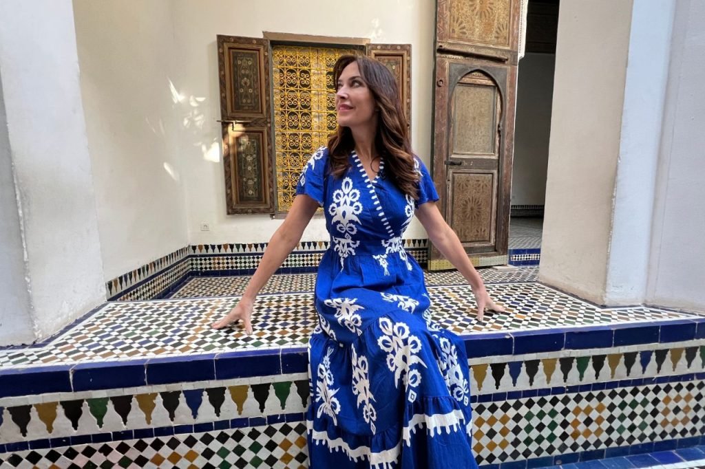 Karen in a blue embroidered dress sitting inside a traditional Moroccan riad with intricate tilework and carved wooden panels.