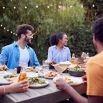 Multi-Cultural Friends At Home Sitting At Table Enjoying Food At Summer Garden Party