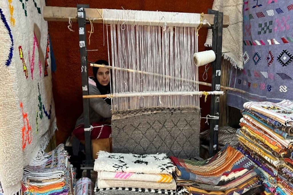 Berber woman weaving a traditional rug by hand in a small shop in the Ourika Valley, Morocco, surrounded by stacks of colorful handwoven carpets and textiles.