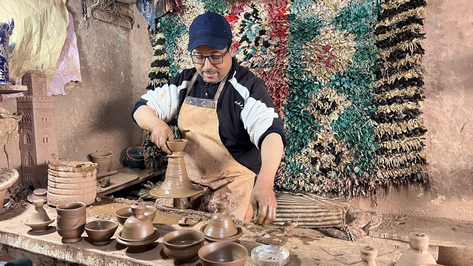 A male artisan in Tafza village shapes clay on a traditional foot-powered pottery wheel, surrounded by freshly made ceramic pieces. He wears an apron and cap, working in a rustic workshop with earthy walls and a colorful woven rug hanging behind him.