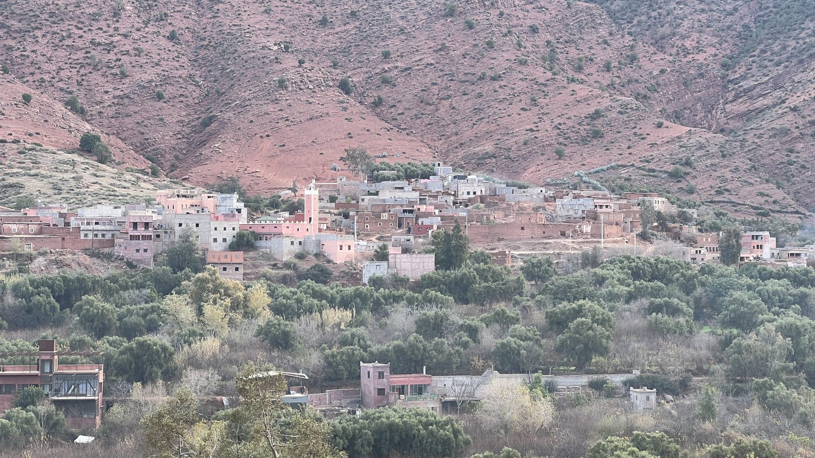 A scenic view of an Amazigh (Berber) village in Morocco’s Ourika Valley, with traditional mud-brick homes nestled against the red mountains and surrounded by lush green farmland.