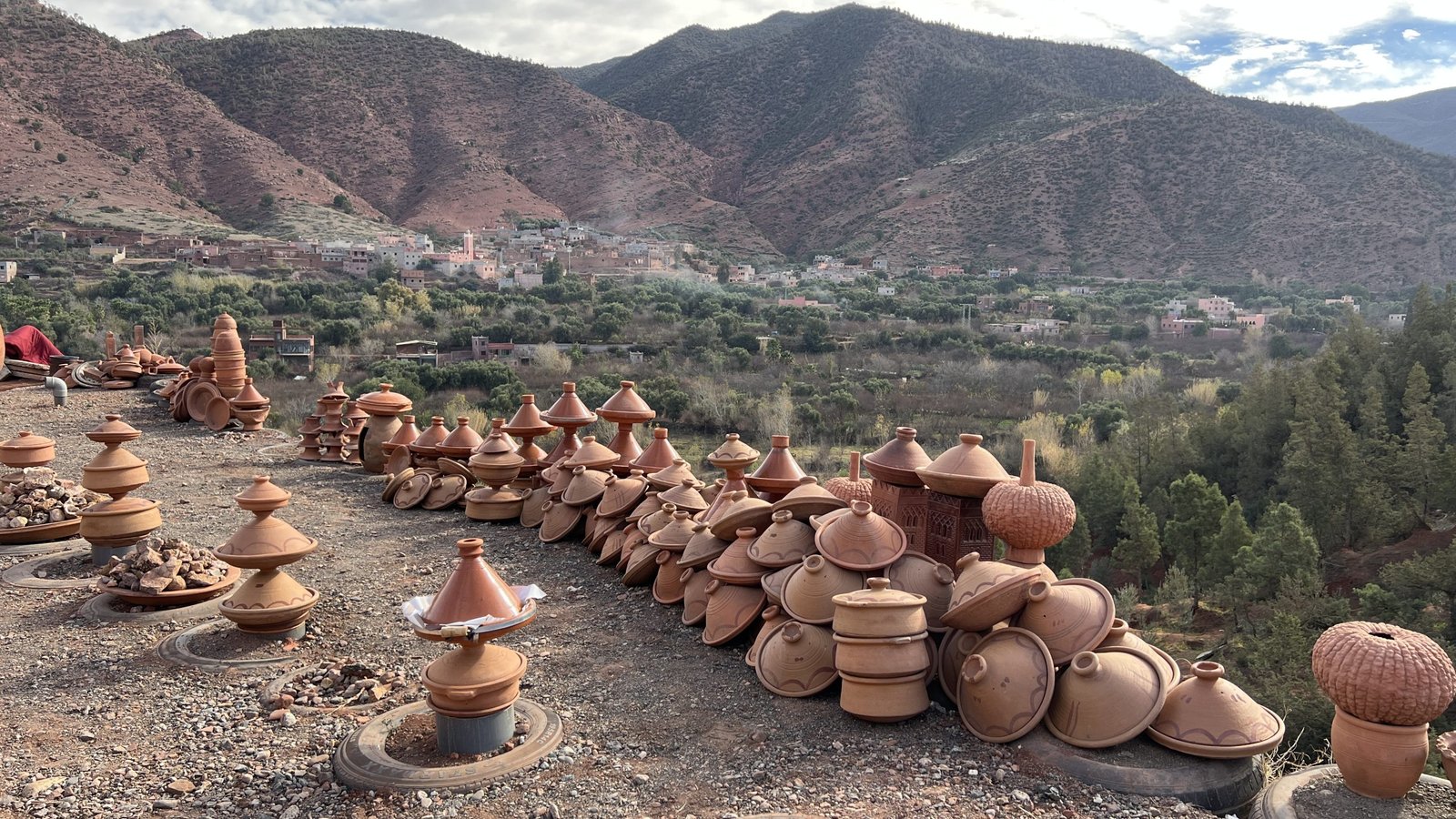 A scenic display of traditional Moroccan pottery, including tagines and clay pots, arranged outdoors on a hillside overlooking the lush green Ourika Valley and the red slopes of the Atlas Mountains.