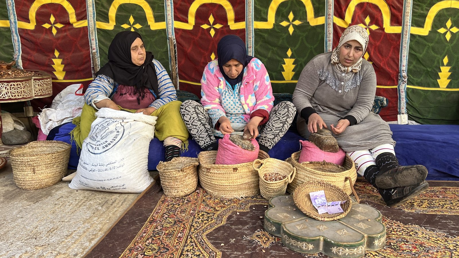 Three Amazigh women sit on cushions inside a traditional tent at Dar Zite, an argan oil cooperative in Sti Fadma, Ourika Valley. They are hand-cracking argan nuts and preparing baskets of seeds, surrounded by woven mats and rustic decor.