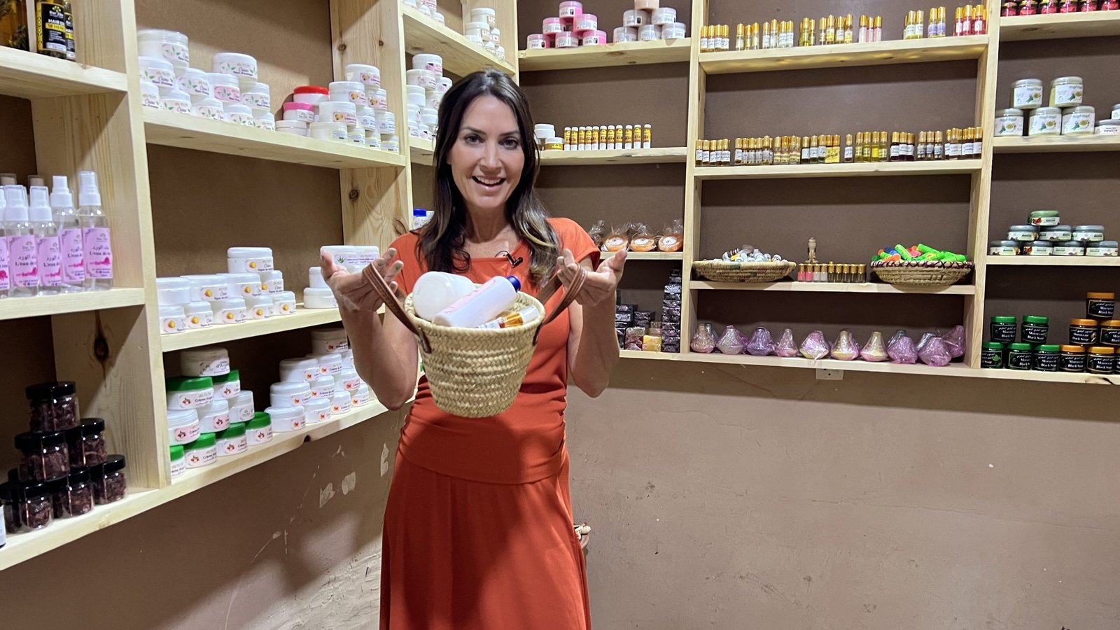 Karen stands inside the Dar Zite cooperative shop in Sti Fadma, smiling and holding a woven basket filled with locally made Moroccan skincare and beauty products. Shelves behind her are lined with argan oil, creams, soaps, and other natural goods.