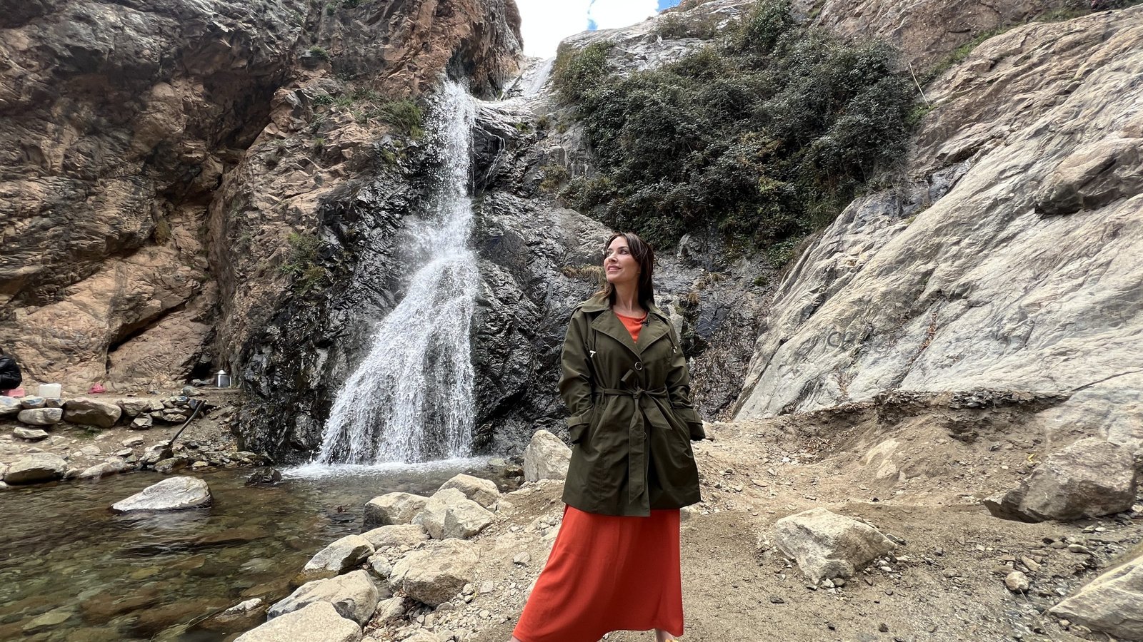 Karen stands near the base of the Setti Fatma Waterfall in Ourika Valley, wearing an olive green jacket over an orange dress. The cascading water flows down rugged rocks into a clear pool surrounded by boulders.