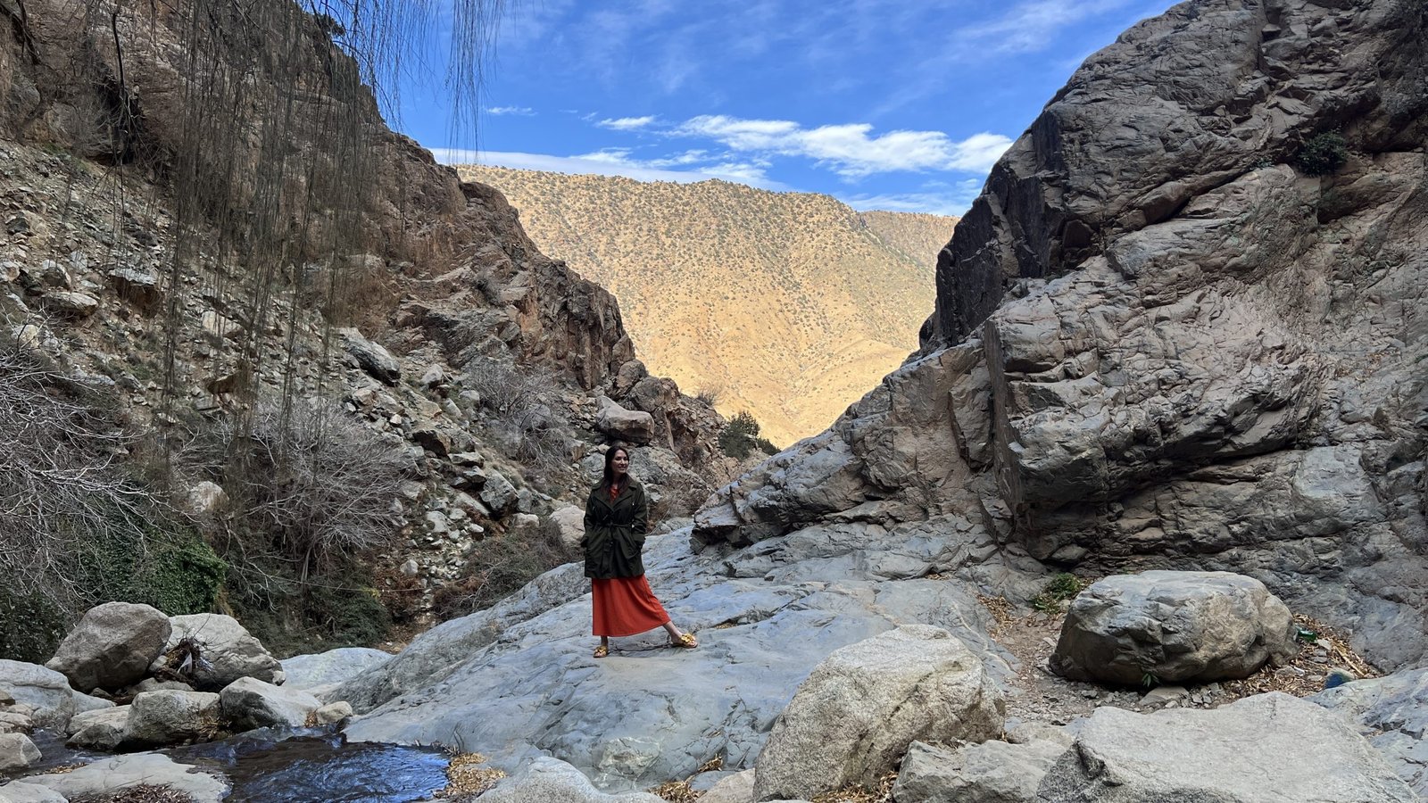 A woman stands on a rocky trail surrounded by dramatic cliffs and mountain views in Morocco’s Ourika Valley, wearing a green jacket over an orange dress. A small stream flows nearby under a clear blue sky.