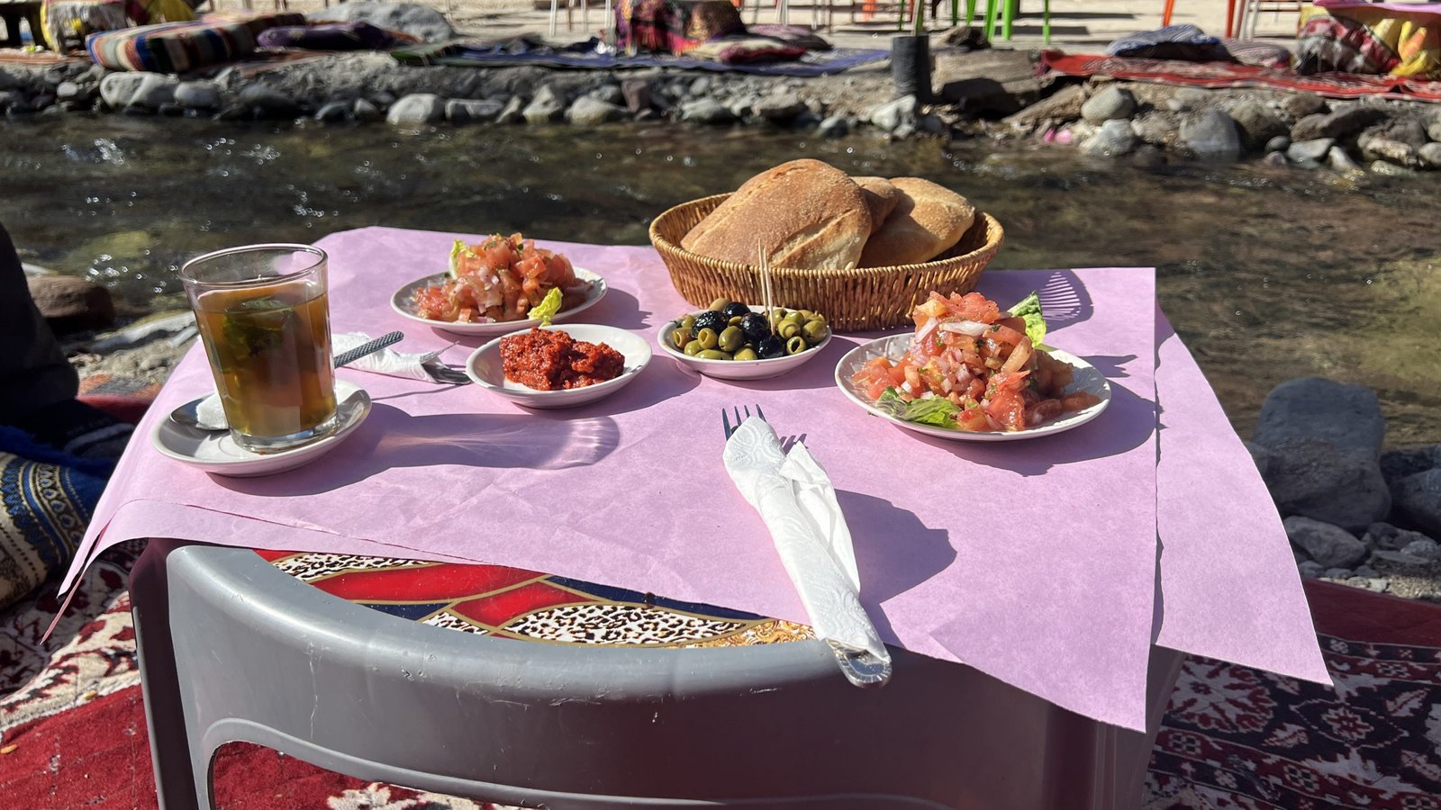 A traditional Moroccan meal served by the riverside in Ourika Valley, featuring fresh tomato salad, olives, spicy harissa, Moroccan bread, and mint tea on a low table set over a rug near the flowing stream.