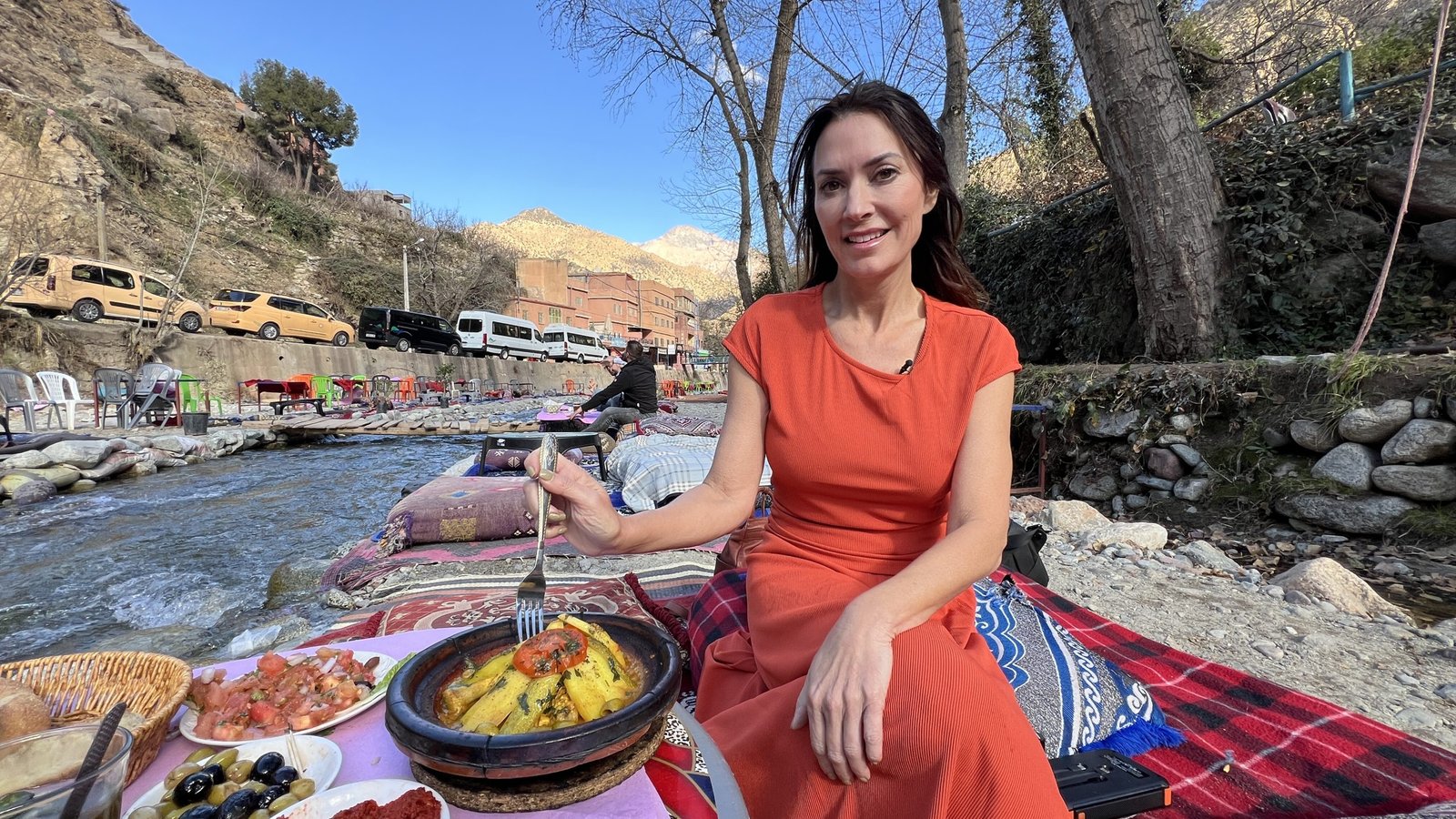 Karen sits on a riverside cushion setup in Setti Fatma, Ourika Valley, enjoying a traditional Moroccan meal with tagine and bread. The river flows beside her, with tables and chairs set up along the banks, and the scenic mountains in the background.