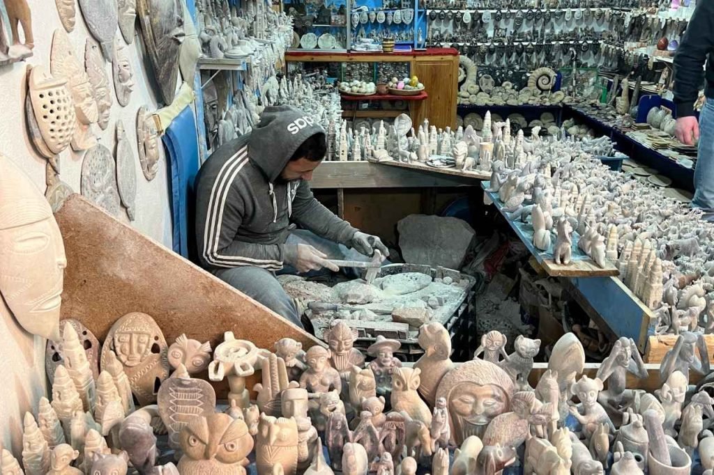 A man in a gray hoodie carves detailed stone sculptures in a small shop filled with intricate handmade figurines and tribal masks in Tafza, Morocco.