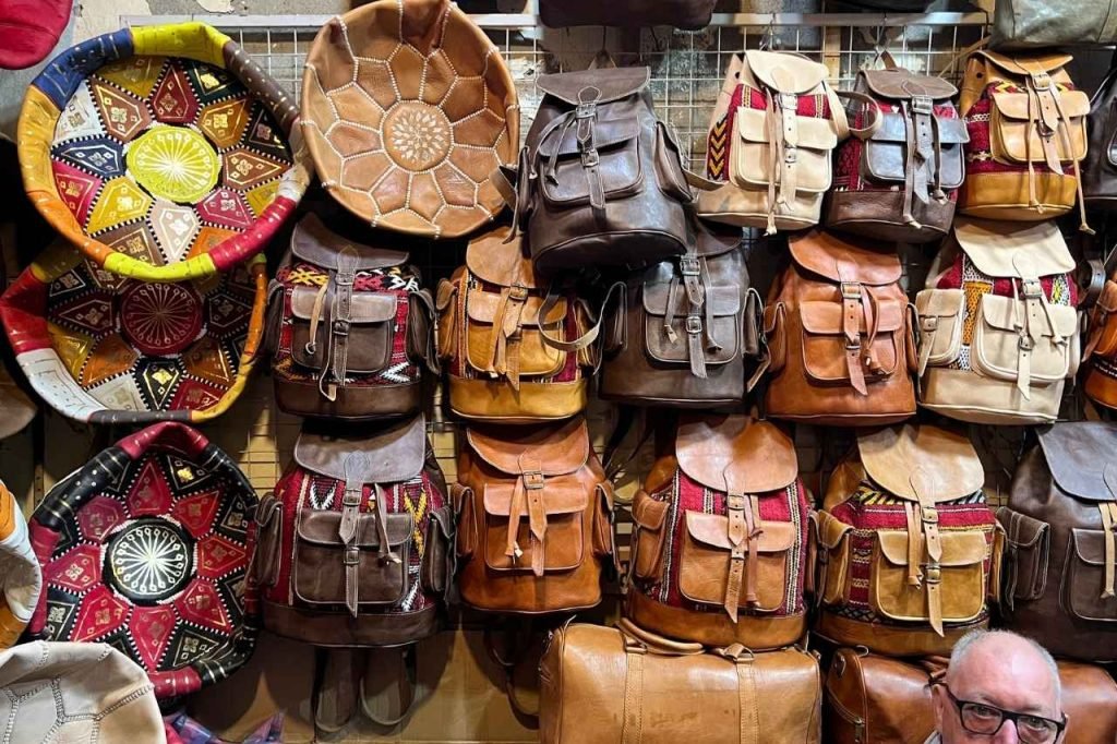 A colorful display of handcrafted leather backpacks and traditional Moroccan poufs for sale at a local market stall in Tnine-de-l’Ourika.