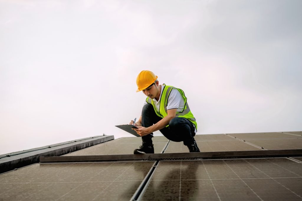 man in a yellow hard hat is writing on a clipboard while kneeling on a roof