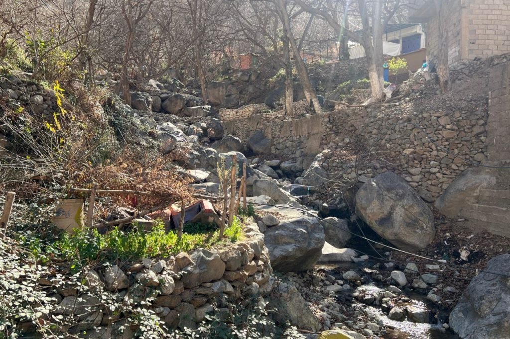 Stone-lined stream running through a rustic Berber village trail in the Ourika Valley, surrounded by dry trees, terraced rock walls, and simple homes.