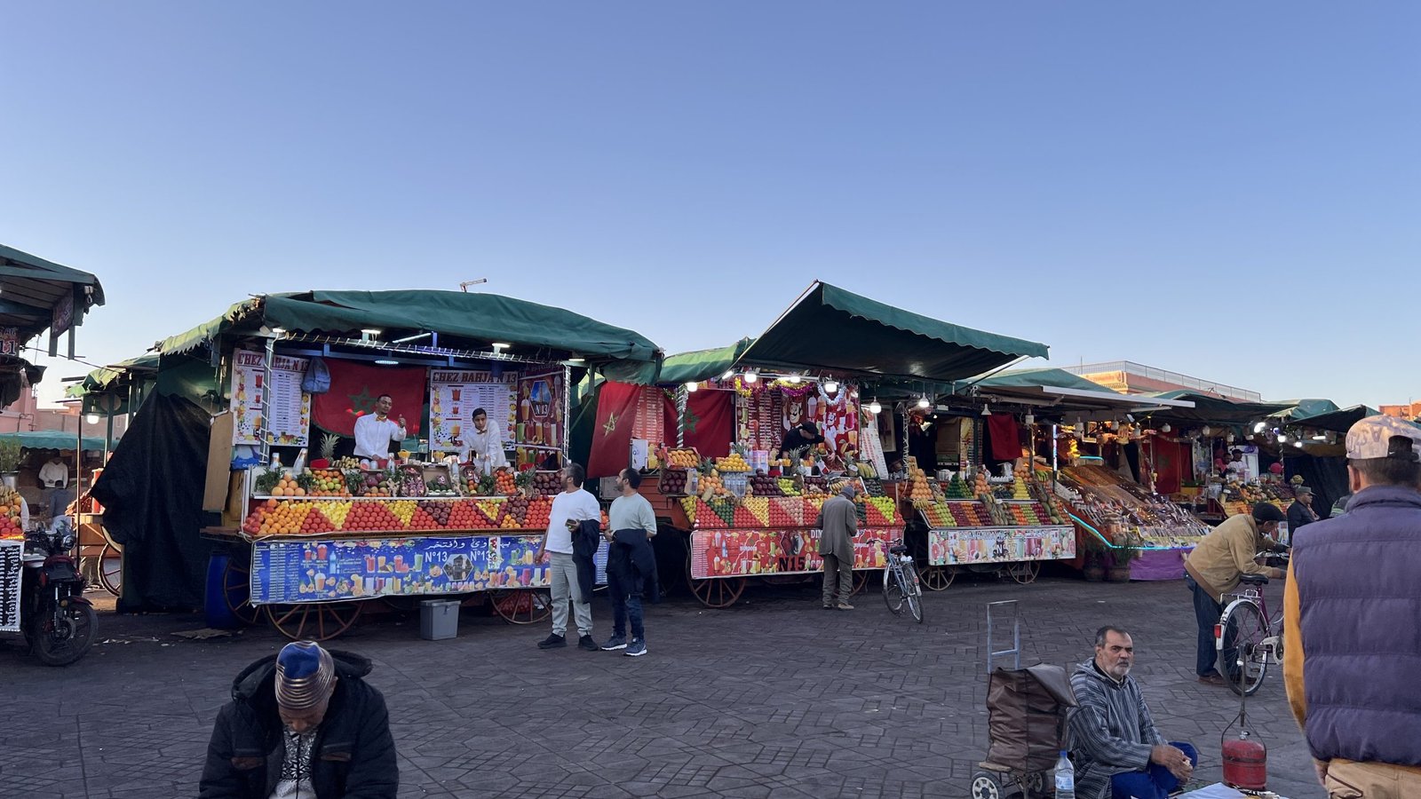 Fruit juice stalls and vendors in Djemaa el-Fna square at sunset in Marrakech, Morocco, with people walking and sitting in the open plaza.