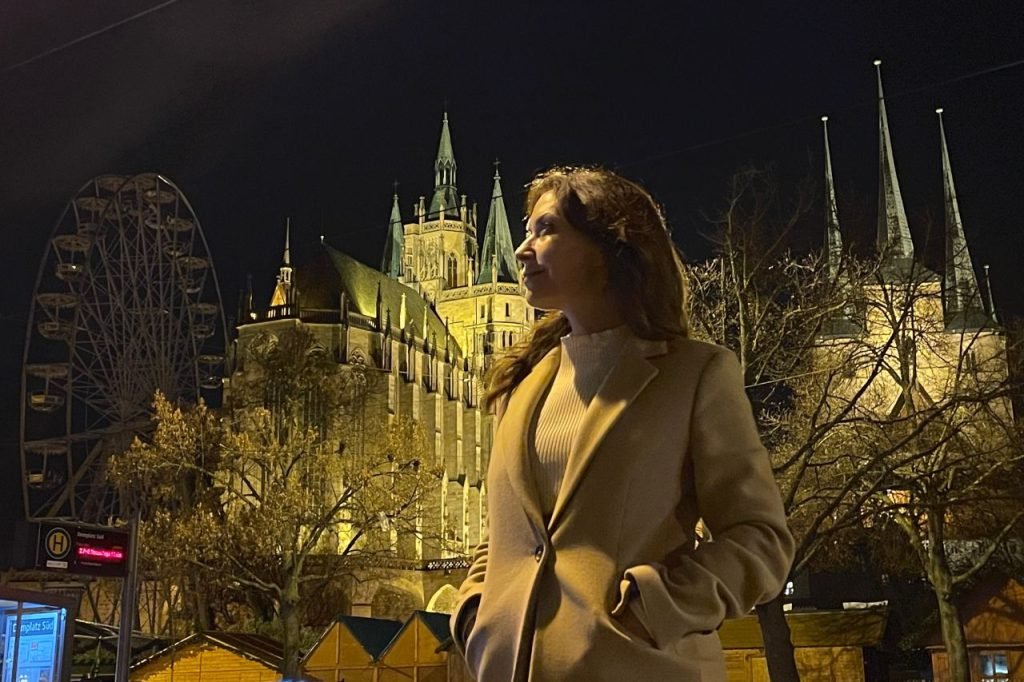 Woman standing in front of the illuminated St. Mary’s Cathedral and St. Severus Church in Erfurt at night, with a Ferris wheel in the background.