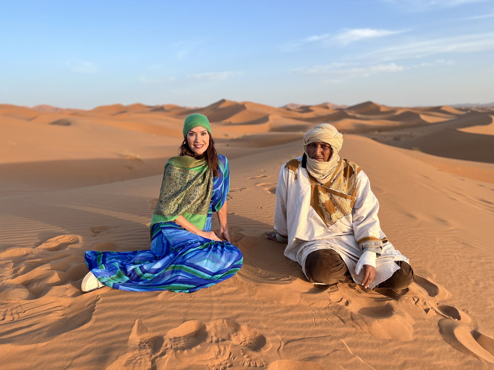 Woman and local guide sitting on the sand dunes near Marrakech, wearing traditional scarves and modest clothing during a 2 day in Marrakech desert visit.