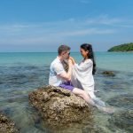 Caucasian man kiss hand of Asian woman also sit on rock in the beach on day time during honey moon