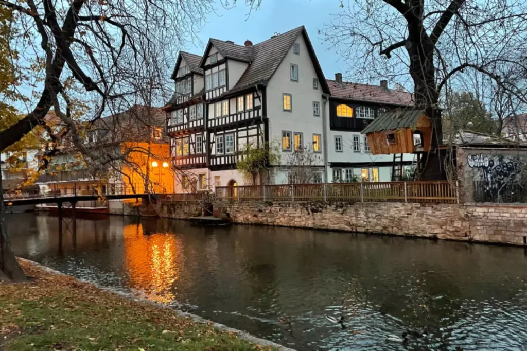 Twilight view of traditional timber-framed houses along the river in Erfurt, Germany, with warm lights reflecting on the water and a small wooden treehouse nestled in a bare-branched tree.