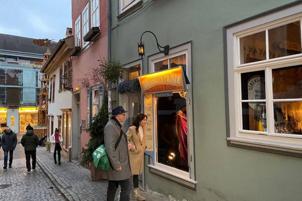 Visitors viewing an illuminated window display of Theatrum Mundi, a mechanical puppet theater on a charming cobblestone street in Erfurt’s old town.