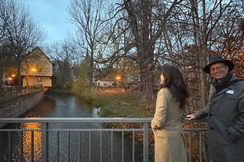 A couple enjoys a peaceful evening view from a bridge over the Gera River, with soft lights reflecting on the water and trees lining the path in Erfurt.