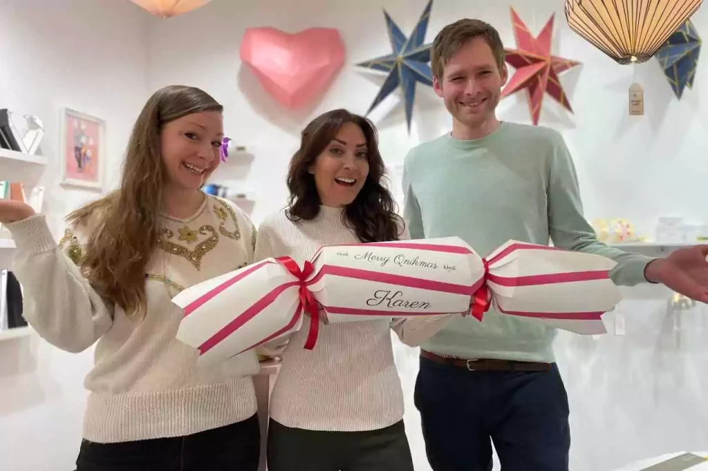 Karen LeBlanc with the founders of Qnik Papier, holding a giant handcrafted paper cracker labeled 'Merry Qnikmas Karen' inside their design-forward paper goods shop in Erfurt, Germany.