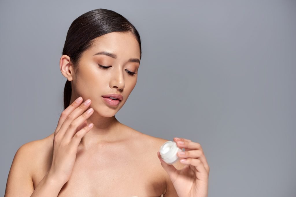 skin care, young asian model with brunette hair holding cosmetic jar and applying face cream