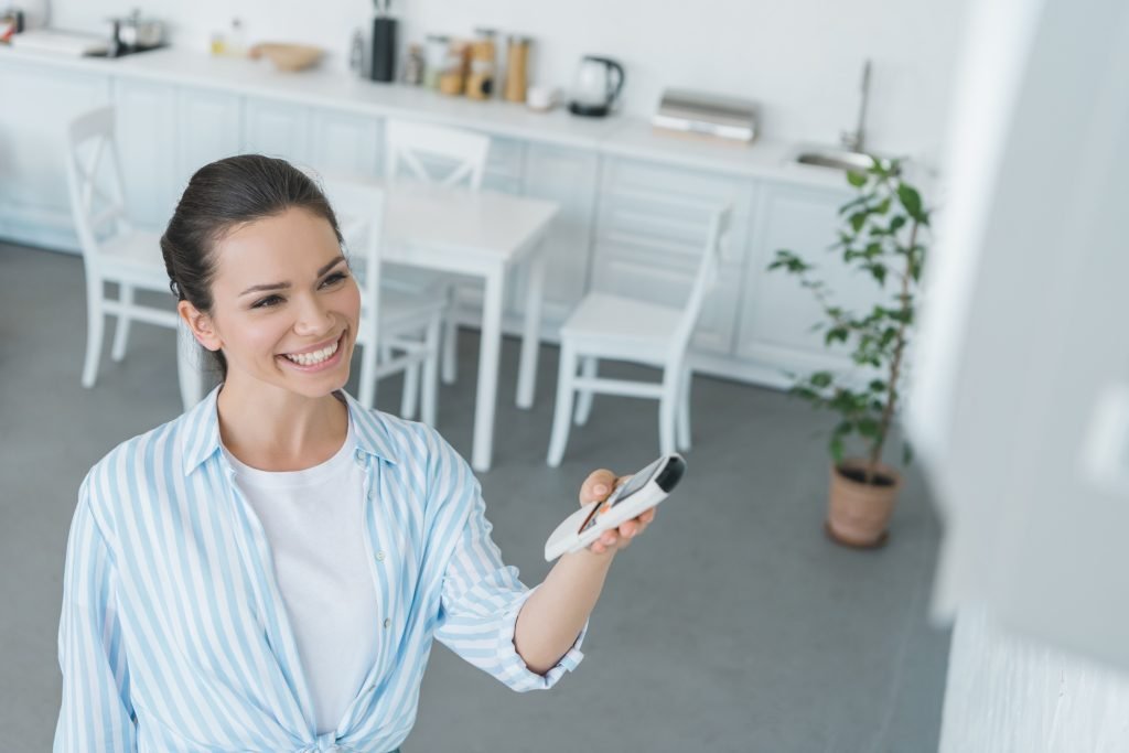 smiling woman turning on air conditioner with remote control at home