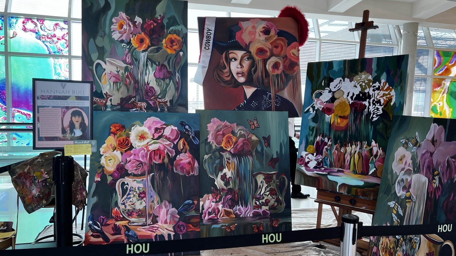 Display of colorful floral paintings by artist Hannah Bull in the Houston Airports Artist in Residence program, with stained glass artwork in the background.