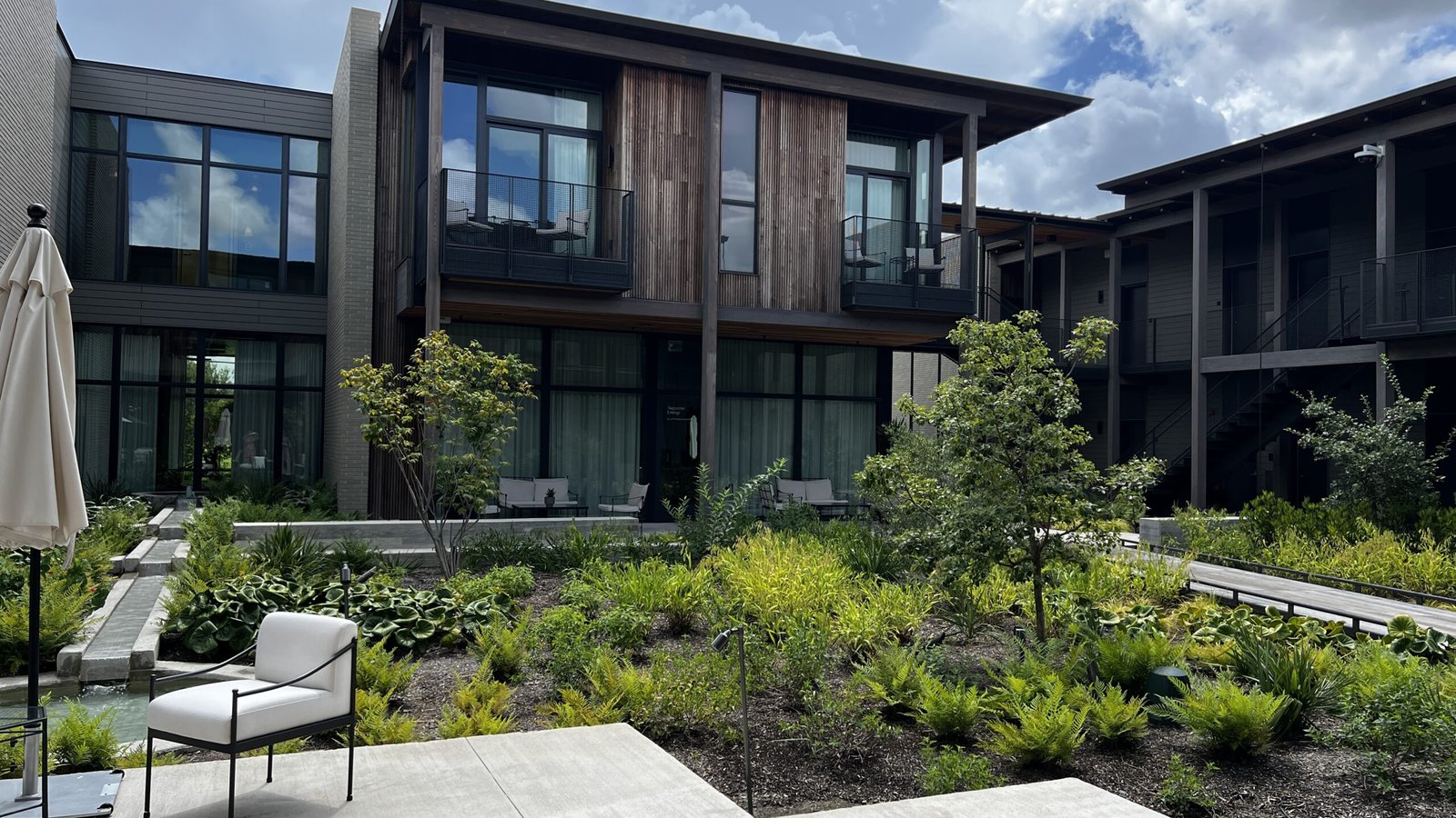 Modern courtyard view of Hotel Saint Augustine Bunkhouse in Houston, with wood and glass facade, landscaped greenery, and outdoor seating.