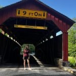 A woman stands in front of the red Lower Brandywine Covered Bridges in Pennsylvania, with a yellow 9-foot clearance sign and a nearby scenic rivers system marker.