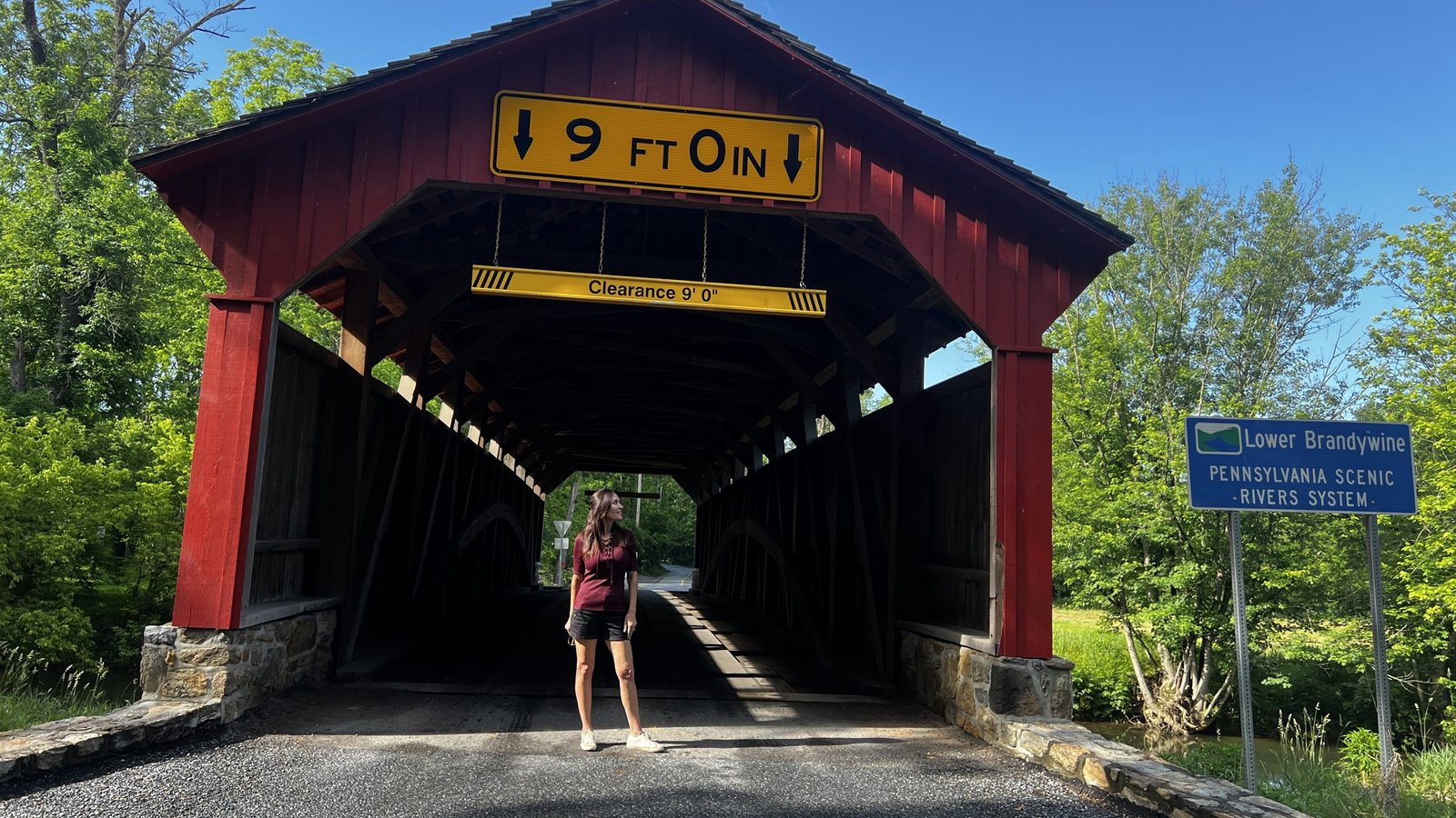 A woman stands in front of the red Lower Brandywine Covered Bridges in Pennsylvania, with a yellow 9-foot clearance sign and a nearby scenic rivers system marker.