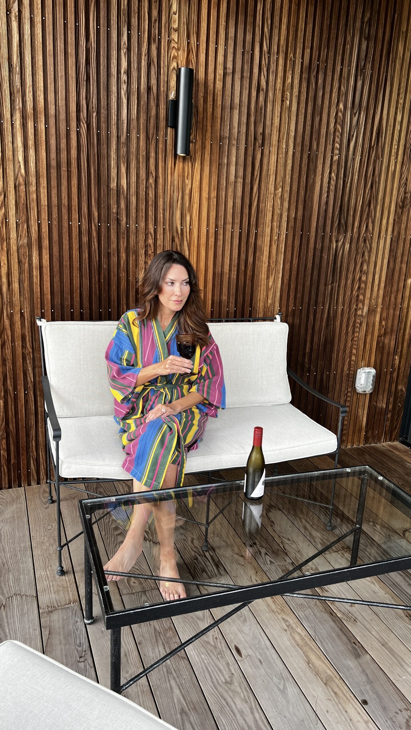 Woman in a colorful striped robe sitting on a white outdoor bench at Hotel Saint Augustine, holding a glass of wine with a wine bottle on the table.