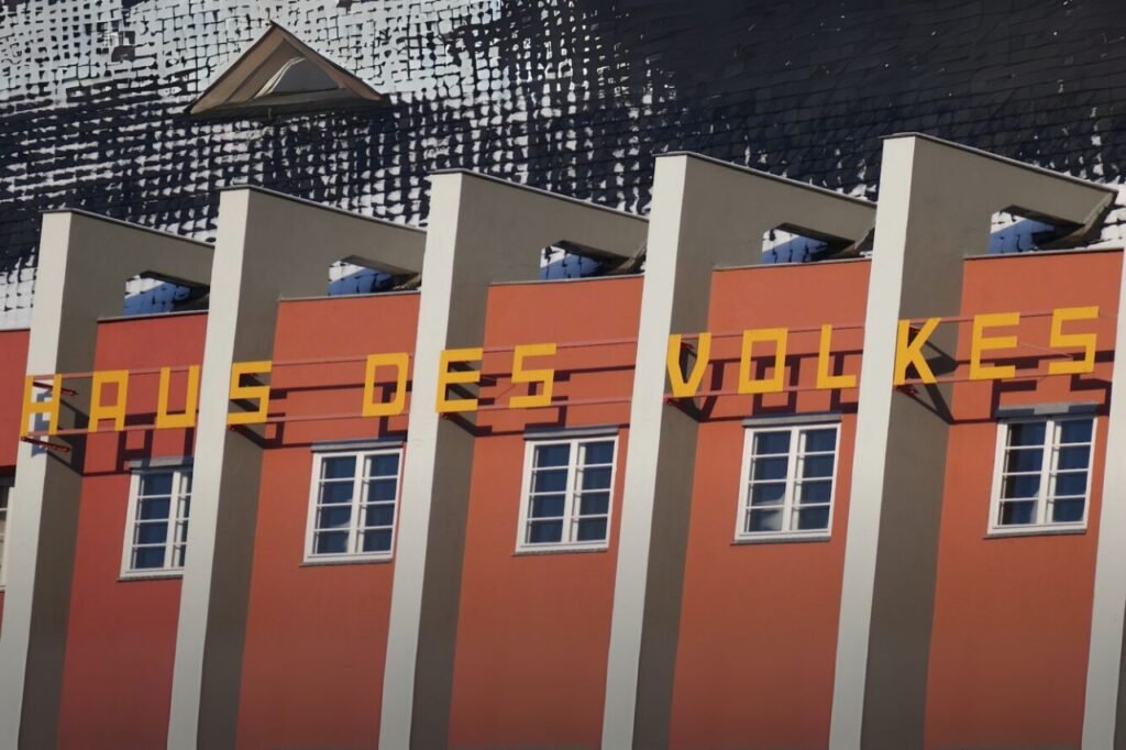 Exterior of the Haus des Volkes in Probstzella, a Bauhaus-designed building with bold lettering and geometric columns against a red and white facade.