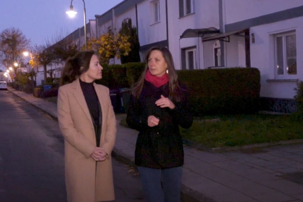 Two women walk and talk through the Törten Housing Settlement in Dessau at dusk, a neighborhood of Bauhaus-designed homes built for factory workers.