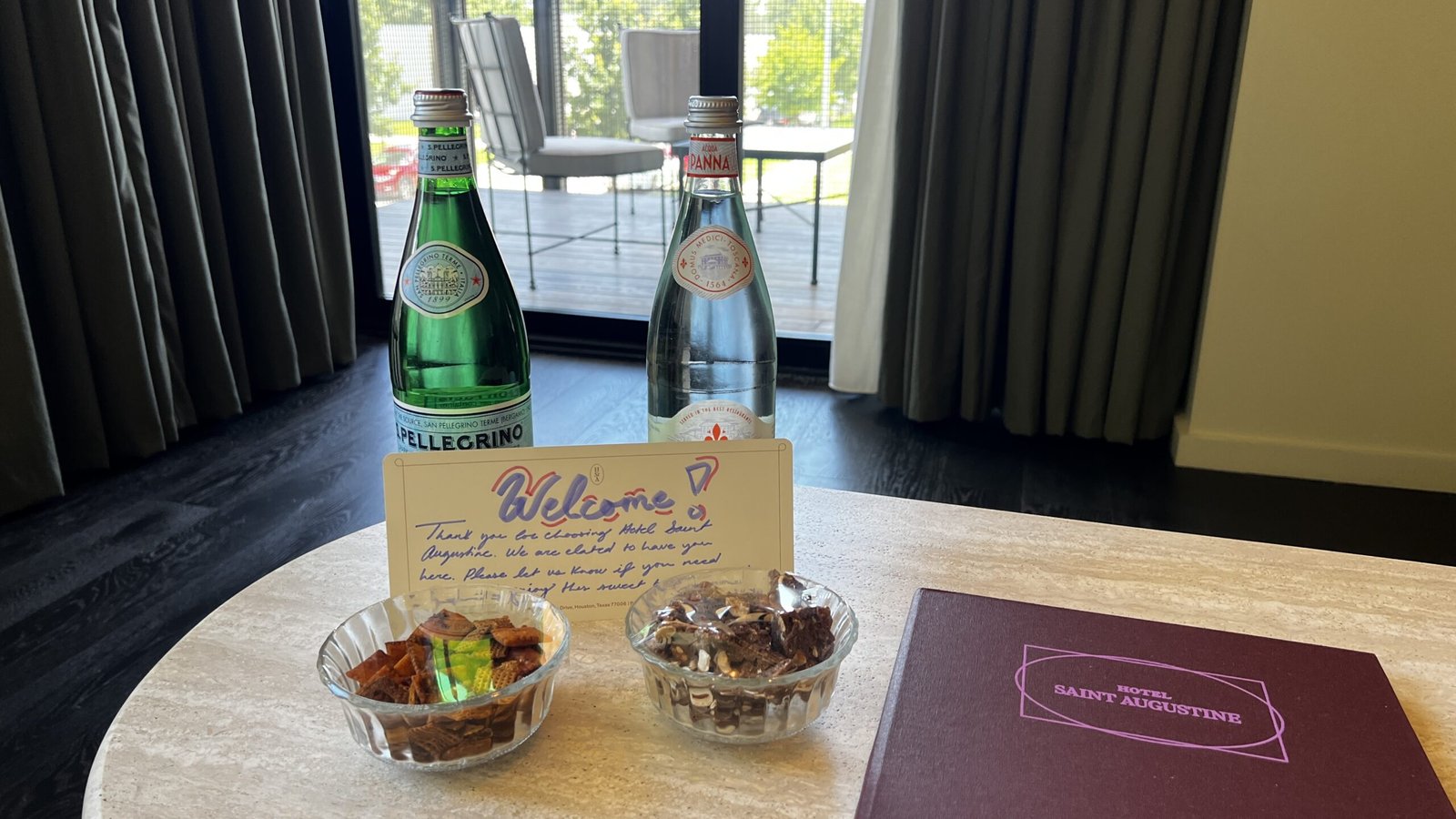 Handwritten welcome note with Pellegrino and Acqua Panna bottles, chocolates, and snacks on a table at Hotel Saint Augustine.