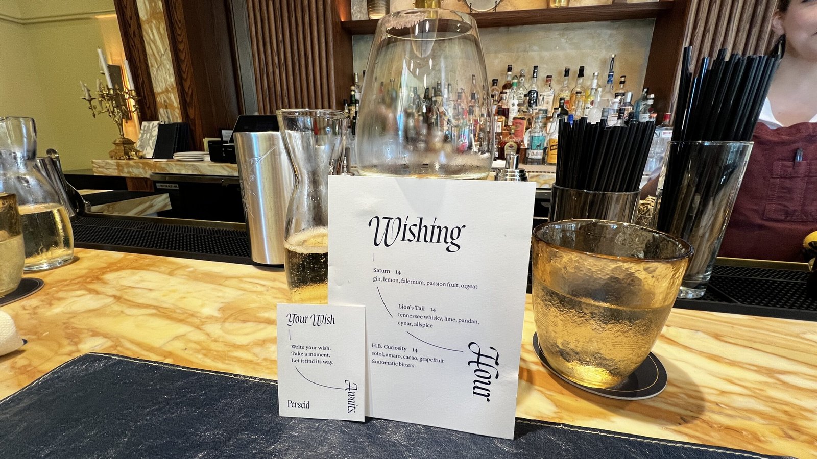 Close-up of Perseid’s Wishing Hour cocktail menu with drinks and golden glassware on the bar counter.