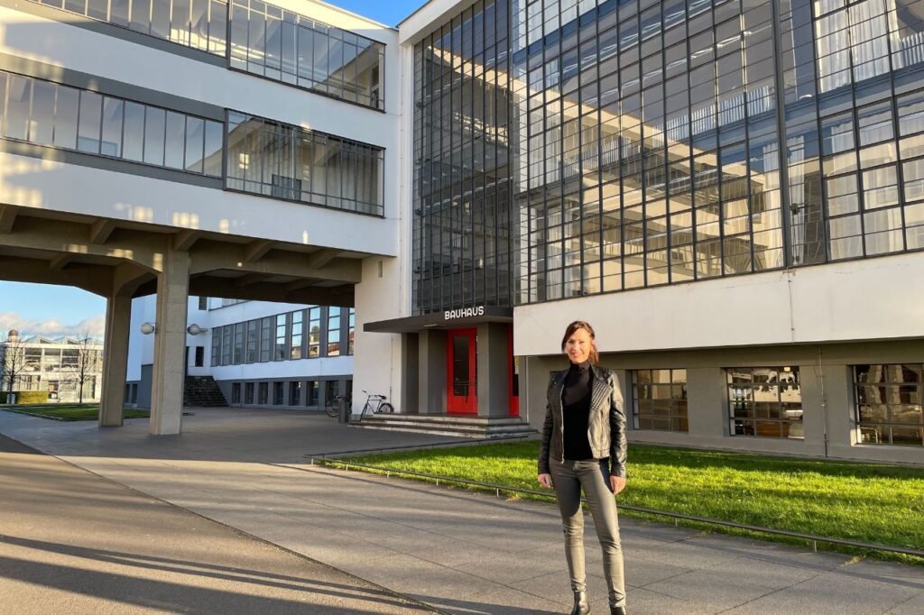 A woman stands outside the iconic Bauhaus School building in Dessau, framed by modernist glass walls and the bold red entrance doors.