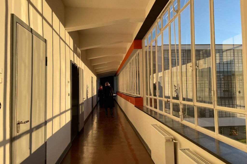 Interior view of a glass hallway in the Bauhaus School in Dessau, Germany, bathed in warm afternoon light with long window shadows across the floor.