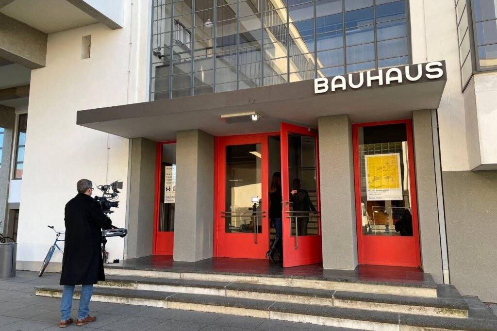 A cameraman films two women entering the iconic Bauhaus School in Dessau through its signature red doors beneath the modernist glass facade.