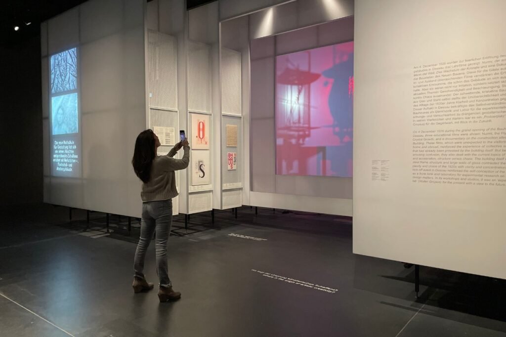 A woman takes a photo inside the Bauhaus Museum Dessau, surrounded by illuminated design exhibits and projected visuals highlighting Bauhaus typography and history.