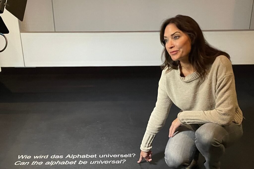 A woman kneels beside text on the museum floor that reads, “Can the alphabet be universal?”—a reflective moment inside the Bauhaus Museum in Dessau.
