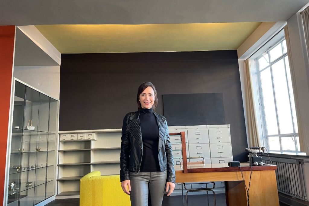 A woman poses inside the recreated Bauhaus director’s office in Dessau, with modernist furniture, a yellow armchair, and natural light filling the space.