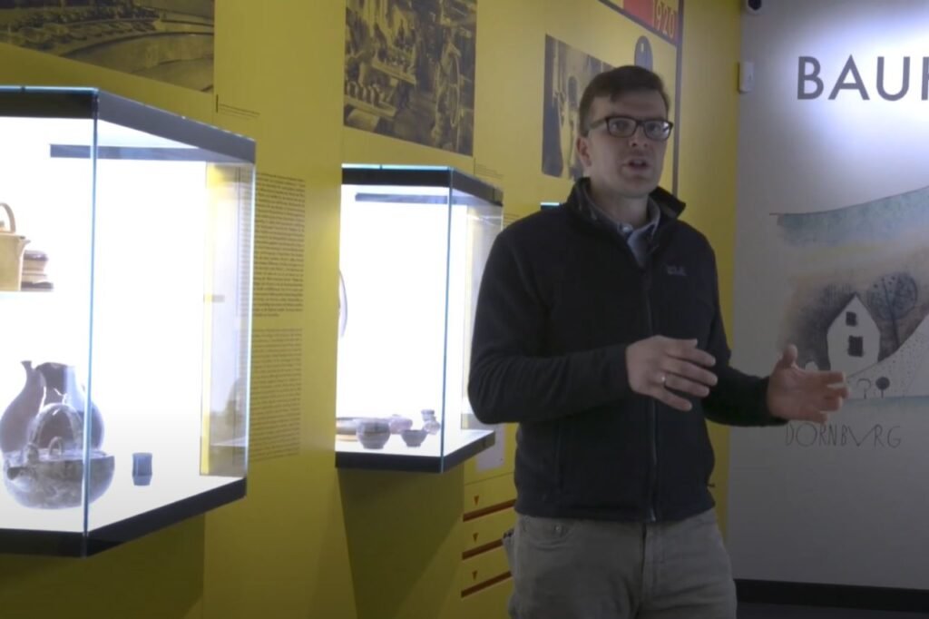 A museum guide stands in front of a yellow exhibit wall at the Dornburg Bauhaus Ceramics Workshop, explaining the historical pottery displays.