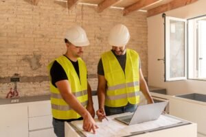 Two contractors wearing safety vests working on laptop and blueprints during a home renovation